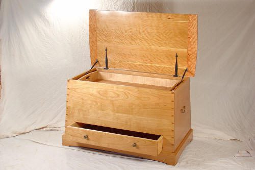 A wooden chest with an open hinged lid and a drawer at the bottom, set against a plain backdrop.