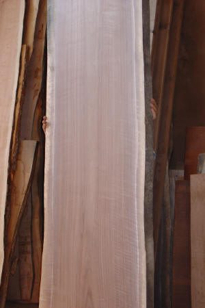 A person is mostly hidden behind a large, vertical wooden plank, with only their hands visible gripping the sides. Other wooden planks are stacked in the background.