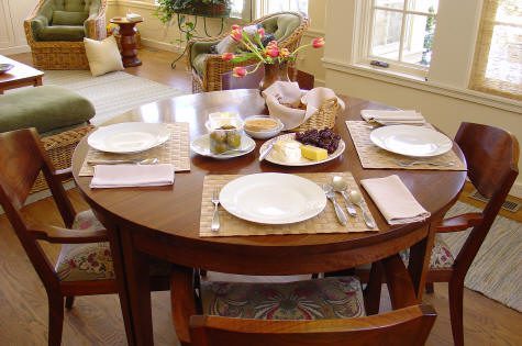 A round wooden dining table set for four with plates, cutlery, napkins, and a selection of food including bread, cheese, grapes, and spreads in a sunlit room.