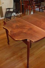 A wooden dining table with a decorative edge and slender legs sits on a hardwood floor, surrounded by other chairs and furniture in the background.