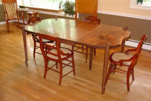 A wooden dining table with four mismatched wooden chairs is placed on a light wood floor in a room with large windows and beige walls.