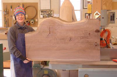 A person wearing a colorful hat and safety headphones holds a large, unfinished wooden slab in a woodworking workshop.