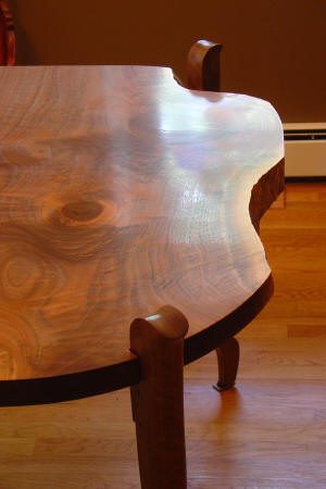 A close-up of a wooden table with a live edge design, showing natural grain patterns and curved legs on a hardwood floor.