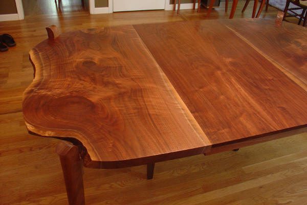 A large wooden table with a natural live edge and rich wood grain sits on a hardwood floor in a dining room.