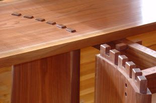 A close-up of a wooden table with exposed dovetail joinery and square inlays on its surface, showcasing detailed woodworking craftsmanship.