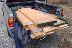 Several large wooden boards are stacked in the bed of a pickup truck with the tailgate down, partially covered by tarps.