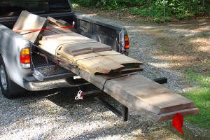A pickup truck with its tailgate down carries long wooden boards secured with straps, extending past the bed with a red flag attached to the end.