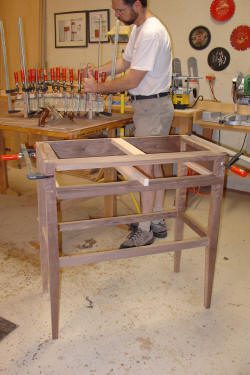 A person assembling a wooden table frame in a workshop, using clamps to hold the frame together. Other woodworking tools and projects are visible in the background.